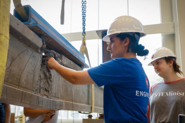 Two women wearing hard hats examine a cement and steel beam suspended from chains.