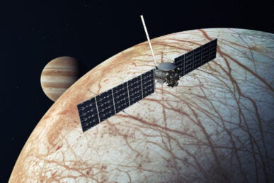 A space craft with two wide rectangular panels hovers above a white and brown moon with the planet Jupiter in the background.