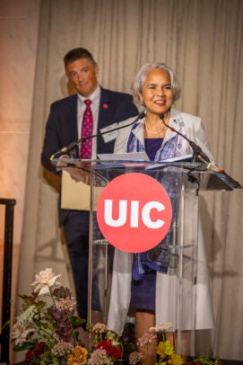 A woman in a grey coat speaks behind a podium