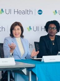 A woman in a blue blazer sits next to a woman in a black suit in front of a repeating graphic that says UI Health with the blue circle UIC logo.