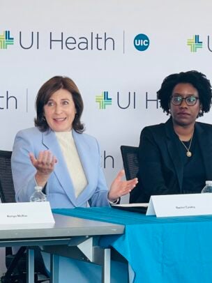 A woman in a blue blazer sits next to a woman in a black suit in front of a repeating graphic that says UI Health with the blue circle UIC logo.