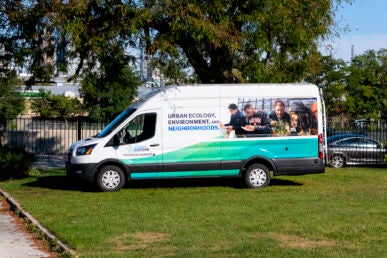 A green and white van with the words "Urban Ecology Environment and Neighborhoods" printed on the side alongside photos of students doing research is parked on grass beneath a tree.