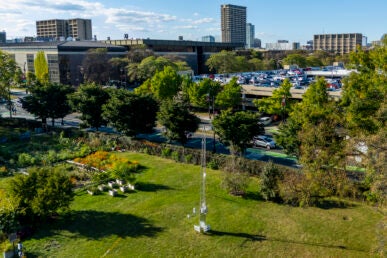 A drone photo of a green field with plants and a tall metal tower in the foreground and campus buildings in the background.