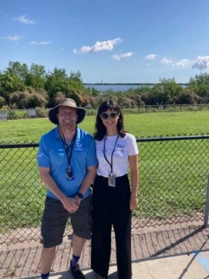 A man and a woman stand in front of metal fence and a green field with the ocean in the background.