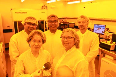 Five people stand in a laboratory with a yellow-tinted light. All five are wearing hair caps and paper gowns.