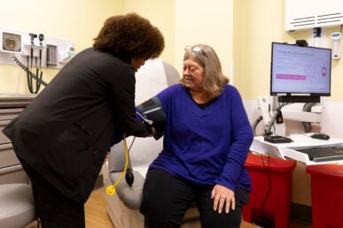 A nurse dressed in black takes the blood pressure of a woman in a purple shirt.