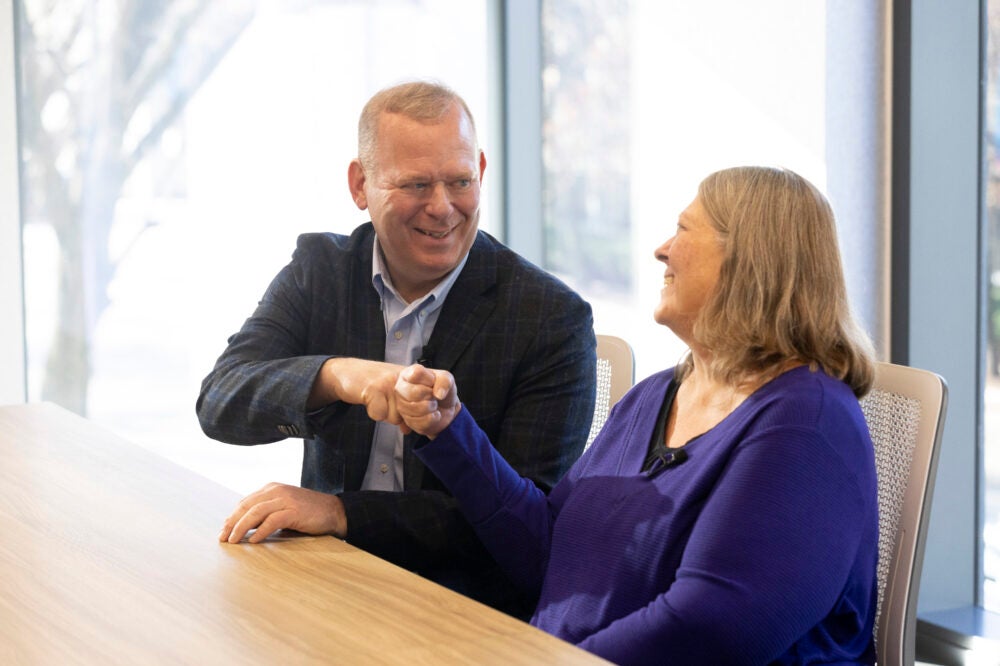 A man in suit jacket and a women in a purple shirt bump fists together.