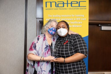 Two women in face masks stand close together holding a glass trophy in front of a yellow vertical banner reading "matec Midwest AIDS Training + Education Center"