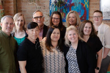 A group photo of ten people posing in front of a red brick wall and multi-colored art.