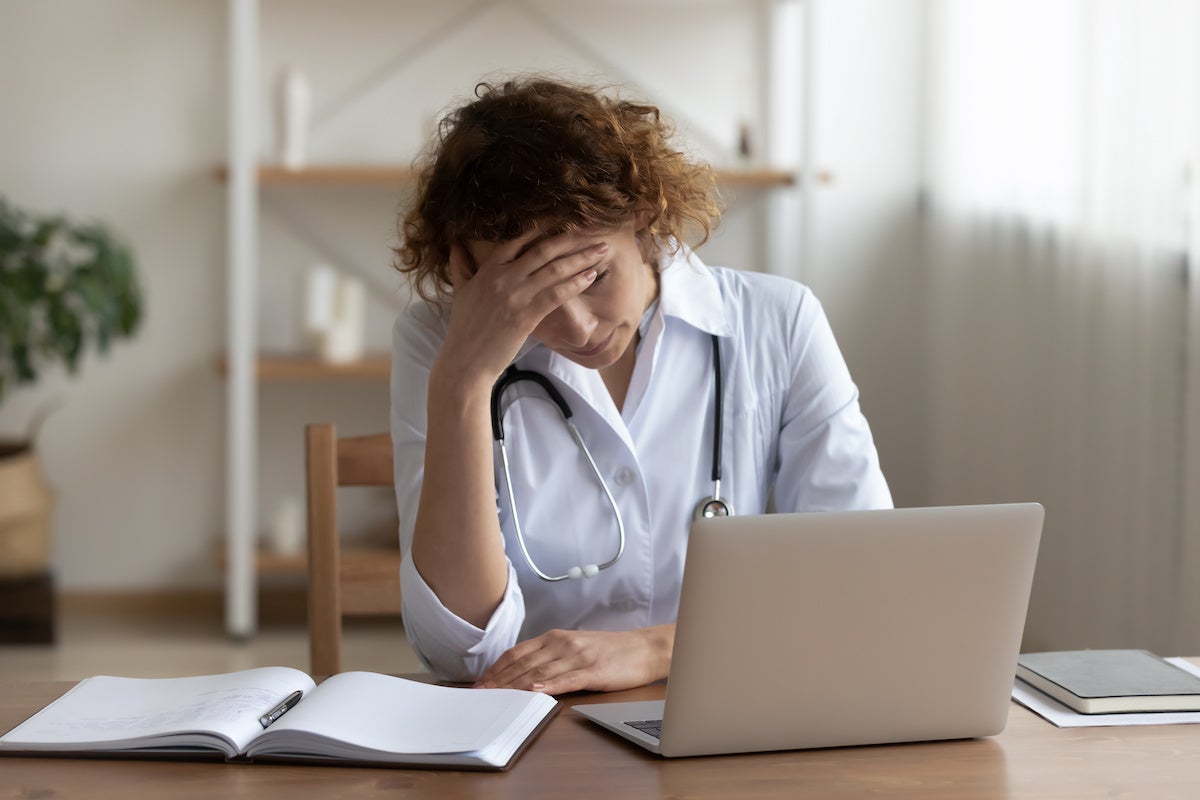 A woman in a white doctor coat with a stethoscope around her neck holds her hand to her forehead while looking at a laptop.