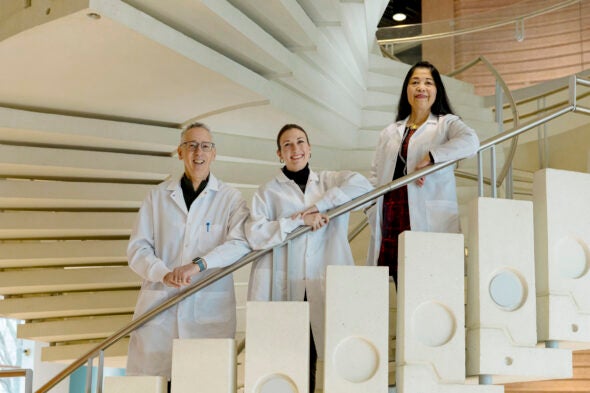 A man and two women in lab coats stand on a white concrete spiral staircase, leaning on a silver railing.