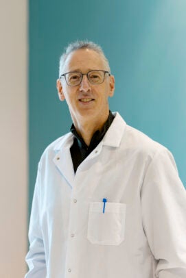 A man in glasses a white lab coat posing in front of a blue wall.