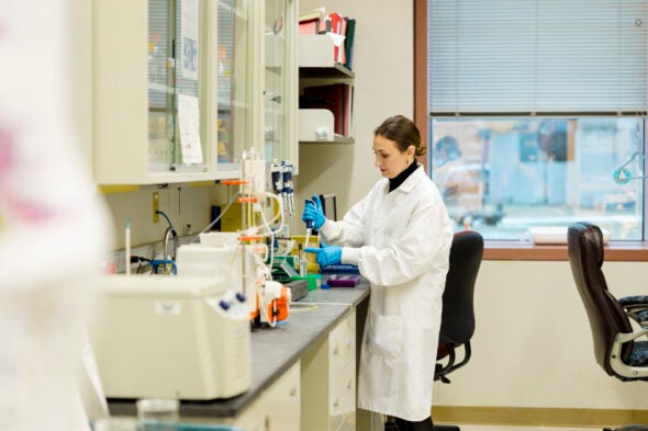 A woman in a white lab coat pipettes at a bench in a laboratory.