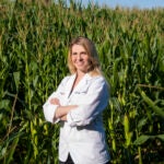 A woman in a white pharmacist coat stands in front of a corn field.