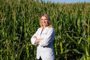 A woman in a white pharmacist coat stands in front of a corn field.