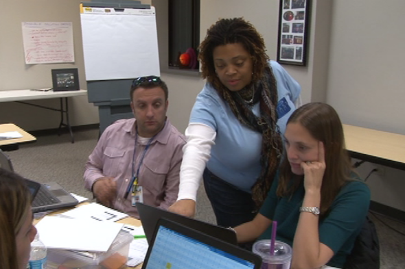 A standing woman in a blue shirt points at a laptop screen as a man and a woman on either side of her look at the screen.